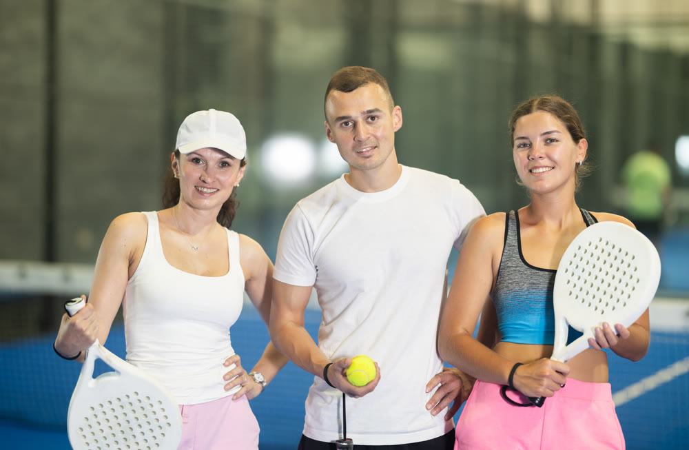 Portrait of three happy athletes - man and two women with padel rackets posing on tennis court