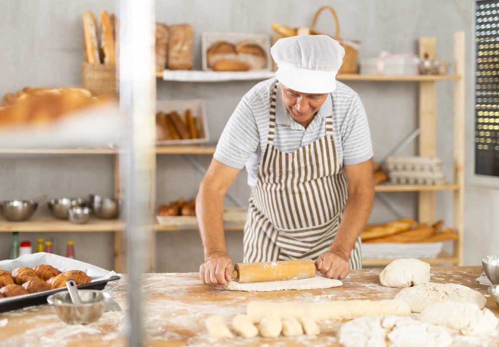 Elderly experienced baker rolls out dough with rolling pin in kitchen