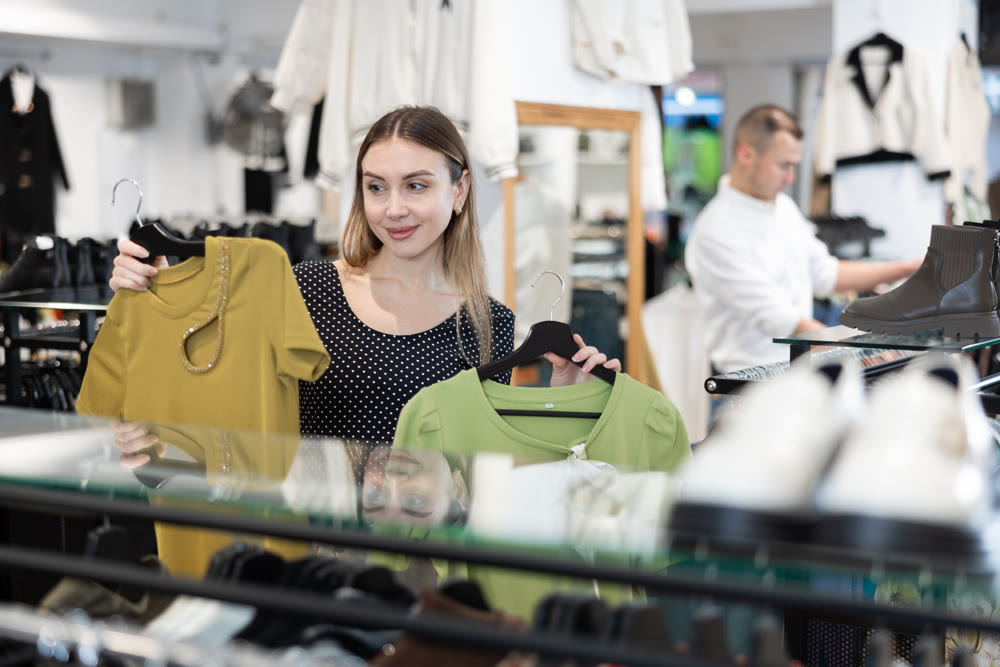 Girl stopped near showcase with outerwear, holds blouse hanger in hands and examines product