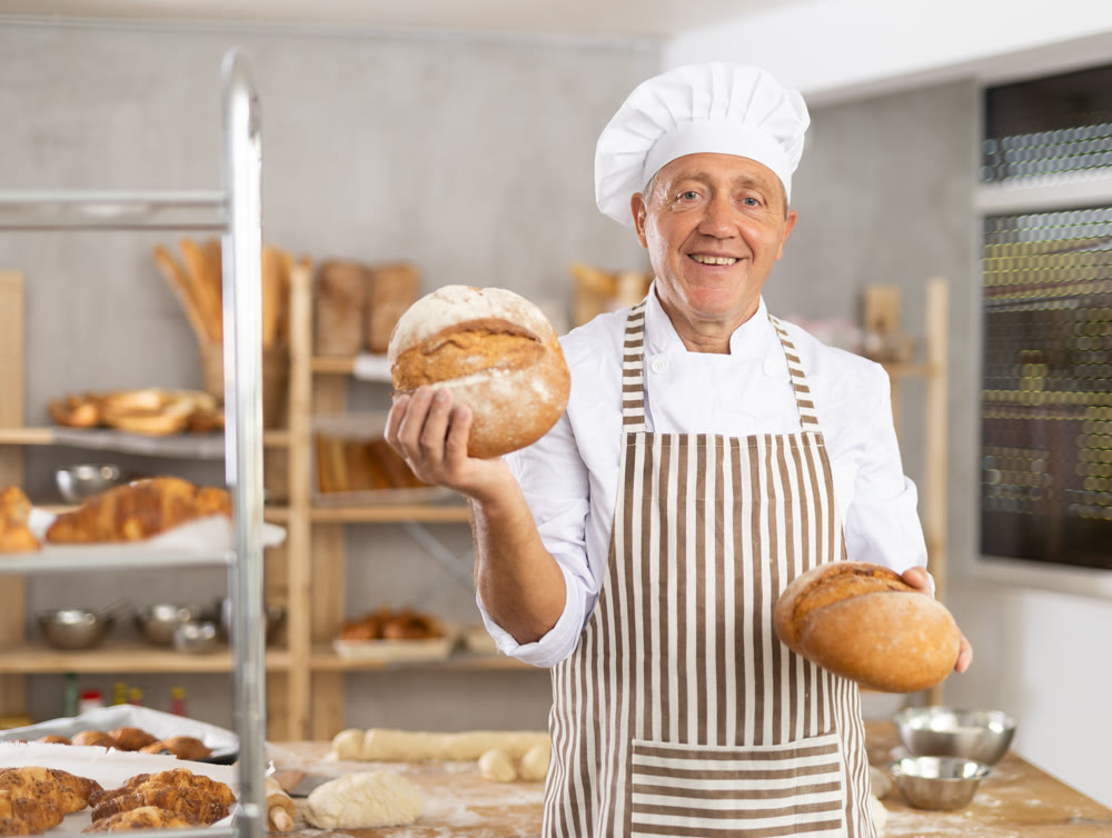 Portrait of elderly baker with bread and baguettes in hands in kitchen