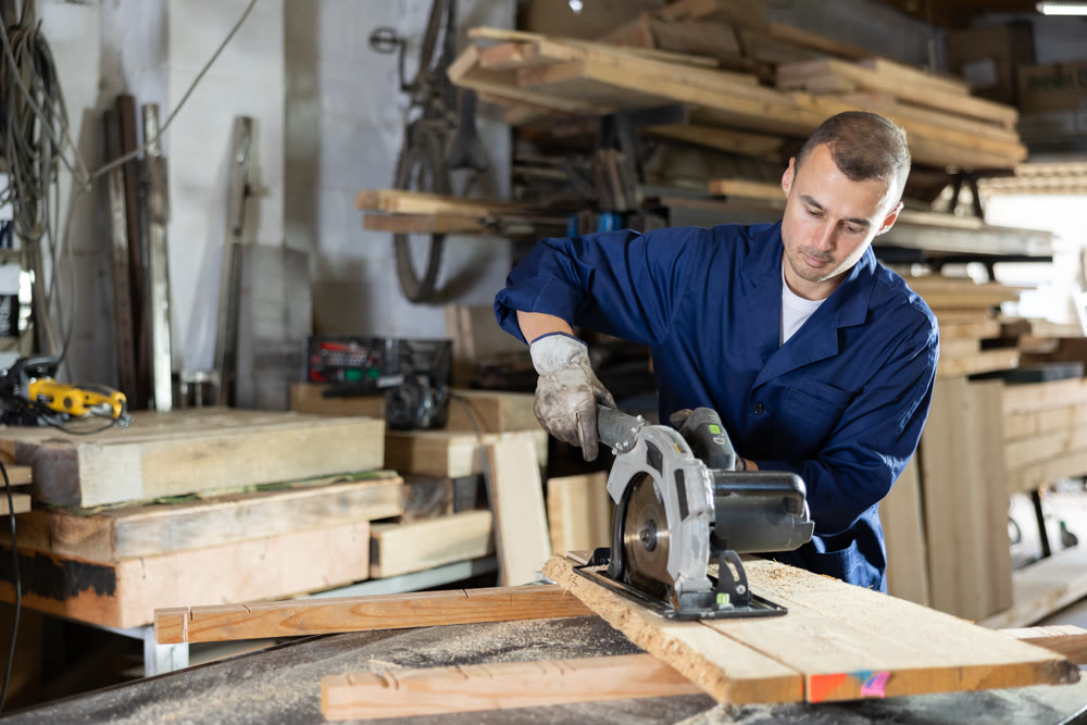 Male employee of workshop cuts wooden board into narrow parts using circular saw