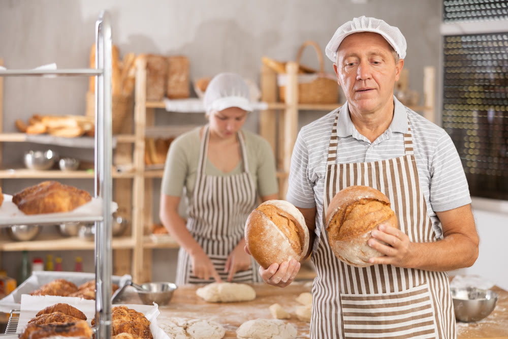 Bakery mature man employee holds ready-made loafs of bread.