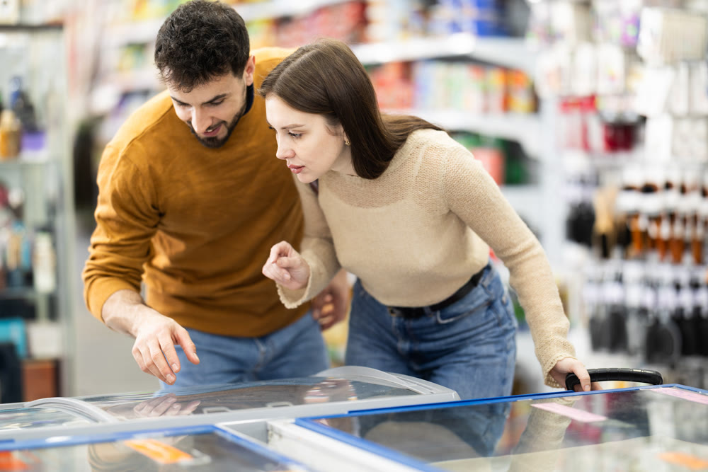 Positive young couple choosing cooled food product standing at fridge in grocery store