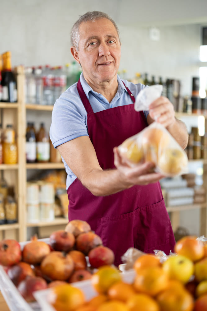 Senior man seller in store is standing near fruit counter, selling apples.