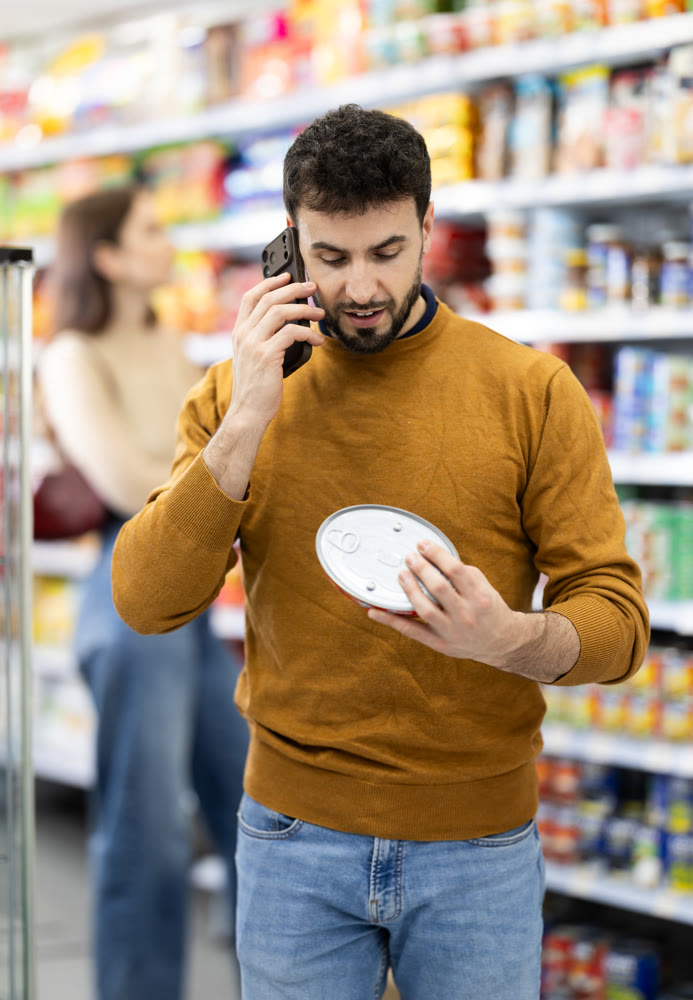 In store, man examines tin can product and talks on phone, advises on purchase.