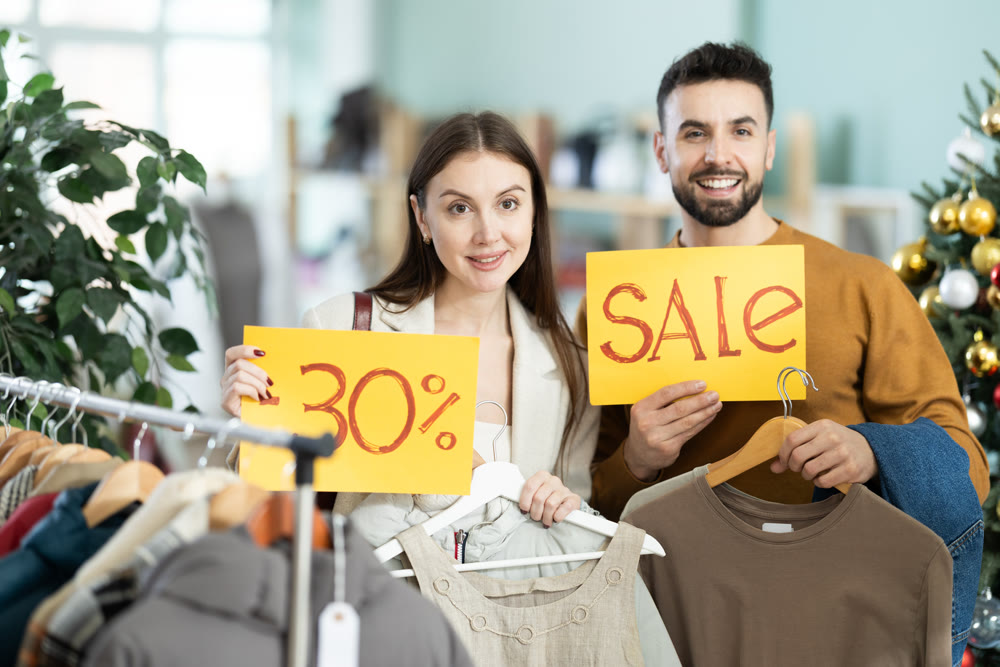 Happy couple, woman and man holding sign saying Sale for winter season standing in interior of clothing boutique
