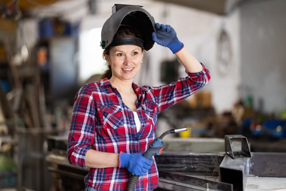 Portrait of a female welder standing with a welding semi-automatic machine and a safety helmet in metal machining workshop
