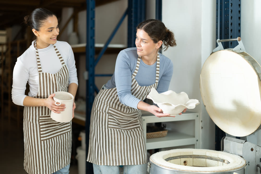 Two female potters take finished ceramic dishes, various mugs and a vase out of oven
