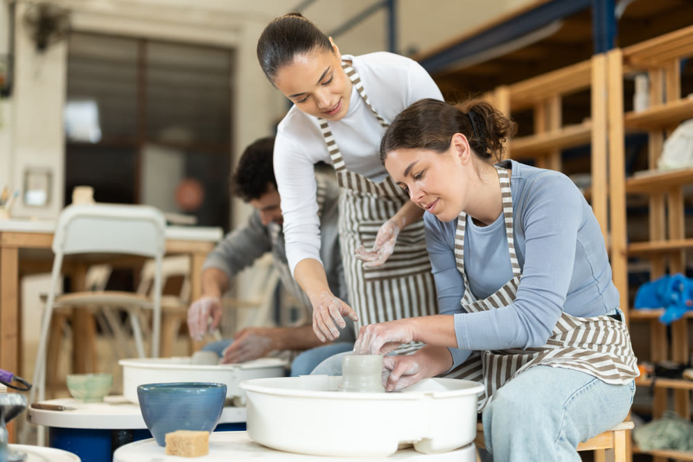 Woman help girl sculpting ceramic product from raw clay on pottery wheel in workshop