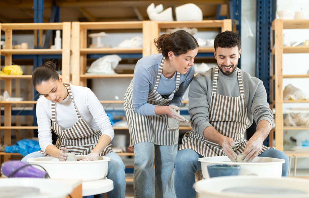 Female teacher help man sculpting ceramic product from clay on pottery wheel in workshop