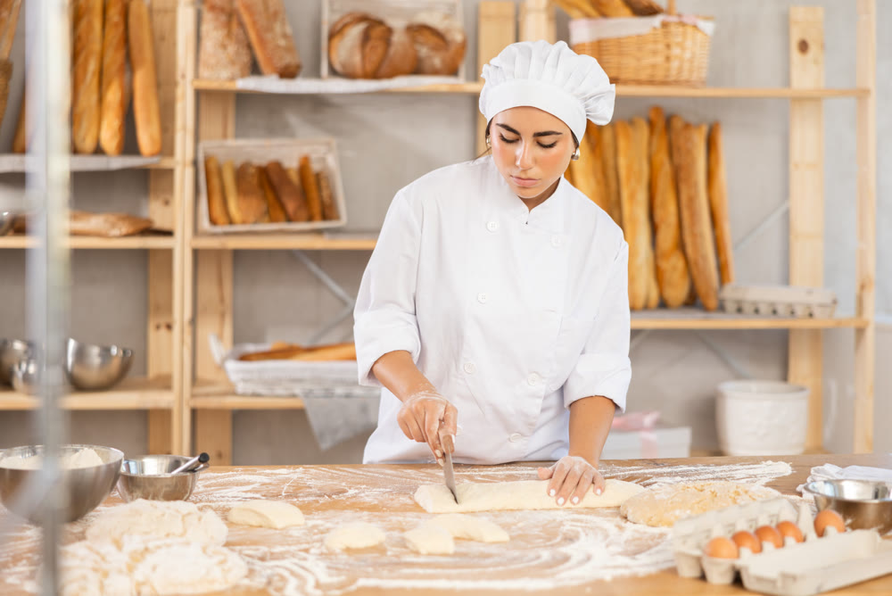 Young female baker cutting dough with scraper