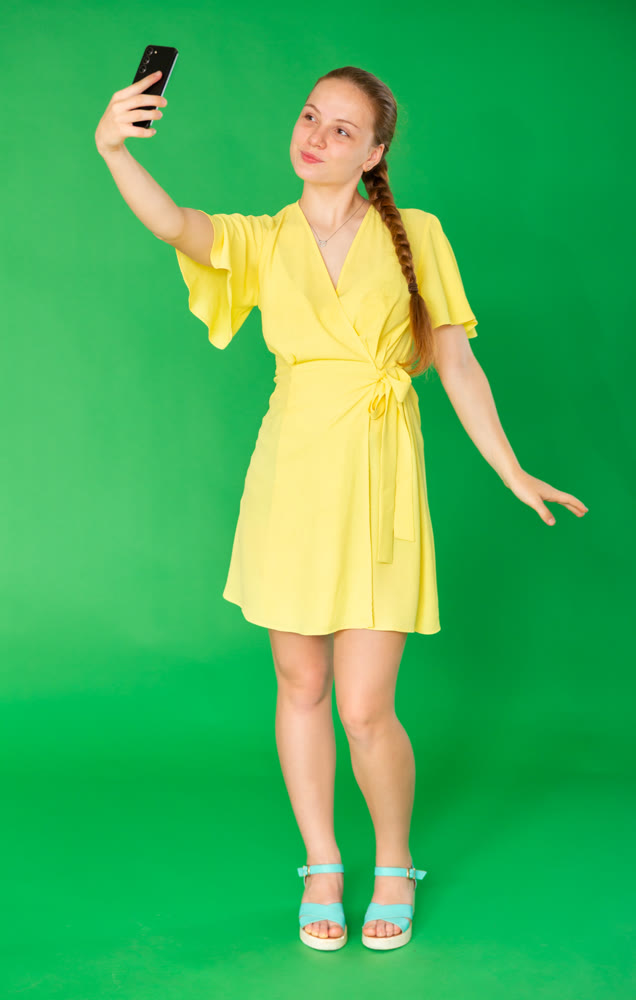 Smiling teenage girl in yellow dress takes selfie on green background. Chroma key compositing.
