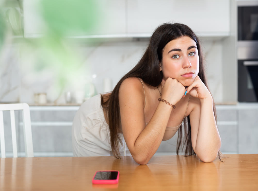 Young woman leaning on kitchen table waiting for call on smartphone