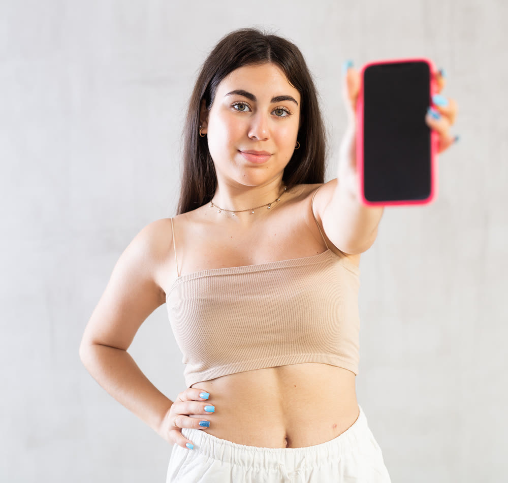 Young woman showing her mobile phone standing against wall