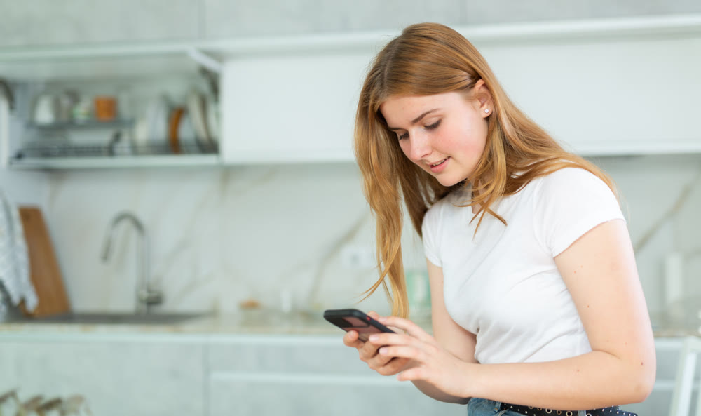 Portrait of focused teen girl in casual wear using mobile phone while surfing Internet in cozy kitchen indoors