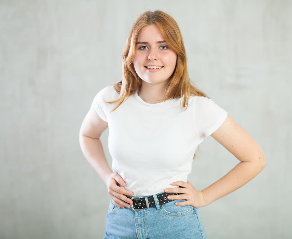 Portrait of positive teen girl in casual wear smiling at camera while posing against concrete gray wall background