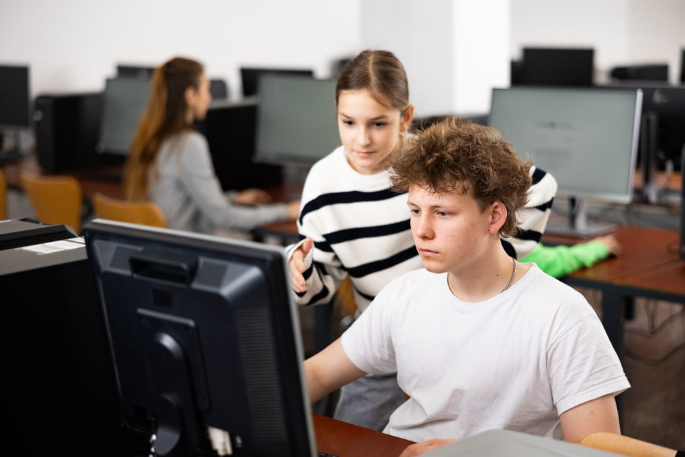 Teen schoolgirl talking to focused classmate in computer class
