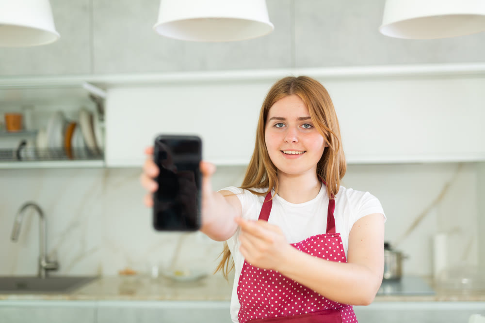 Happy teen girl in red polka dot apron showing modern smartphone with black screen in modern kitchen
