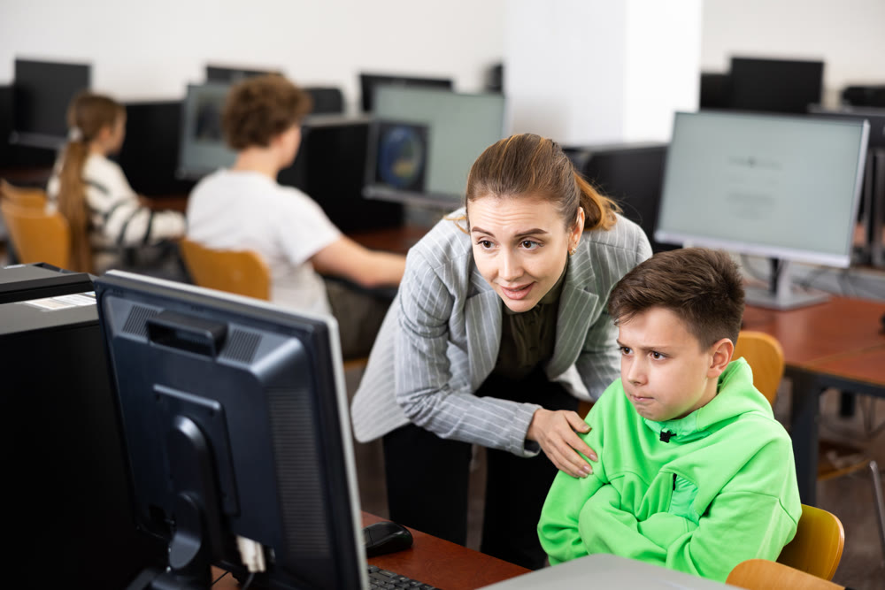 Positive female teacher tells a teenager how to work on computer