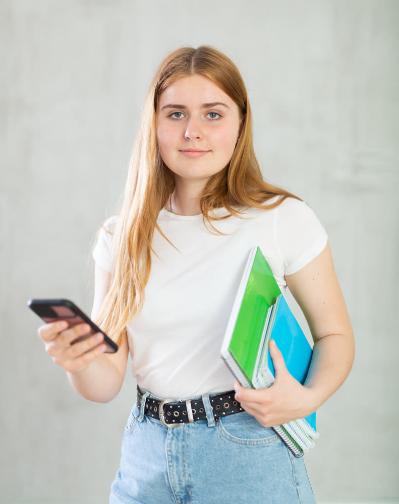 Portrait of focused teen girl in white t-shirt and blue jeans with spiral notepads searching info on smartphone at gray wall. Copy space for advertising