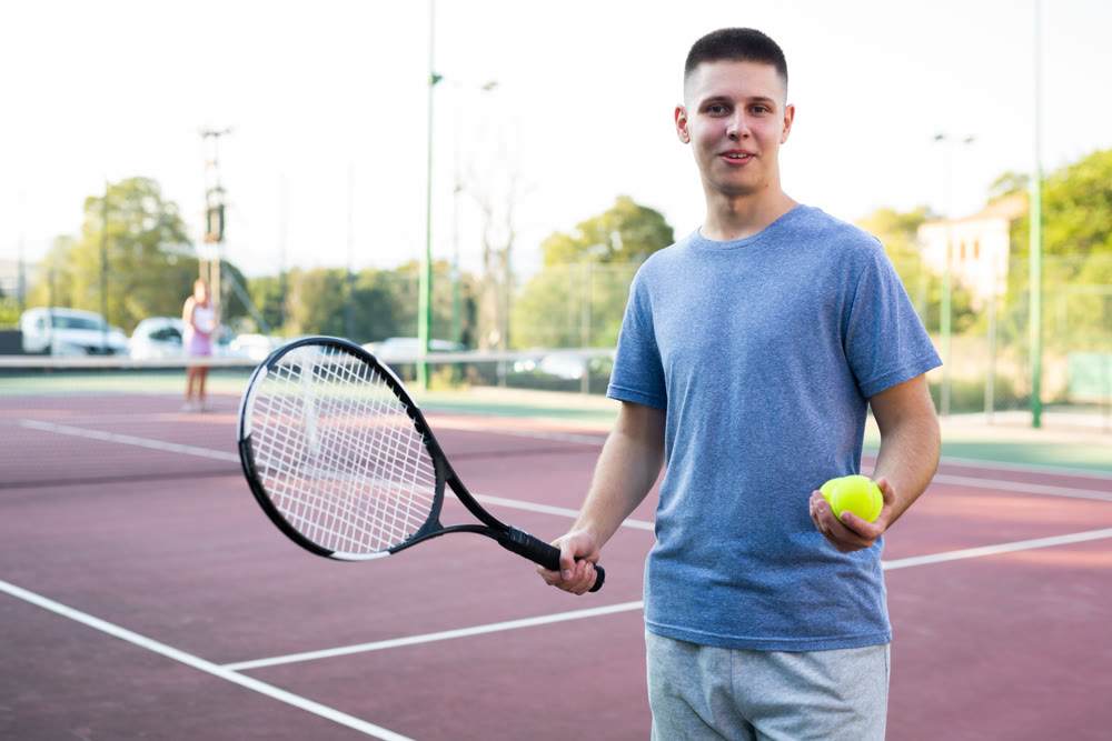 Portrait of positive young man with racket and ball in his hands on tennis court outdoor