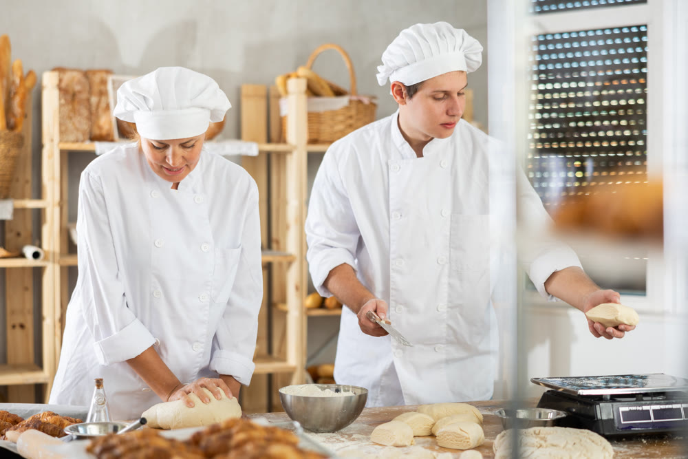 Experienced female adult baker and young male assistant in white uniform kneading dough cutting into pieces and weighing preparing for baking pastries in bakery