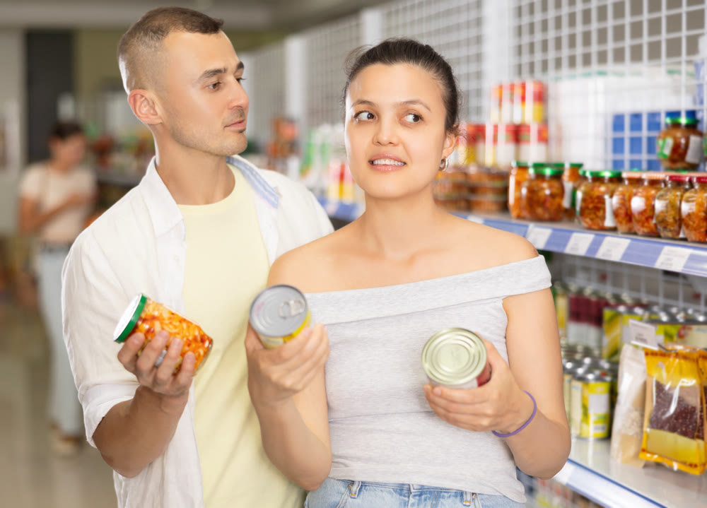 Smiling couple choosing canned vegetables on shelves of supermarket