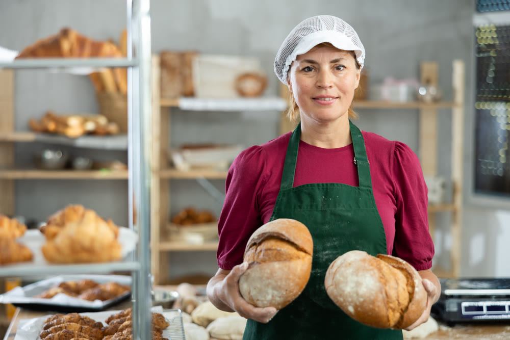 In working room of bakery, there is woman with loafs of bread in hands