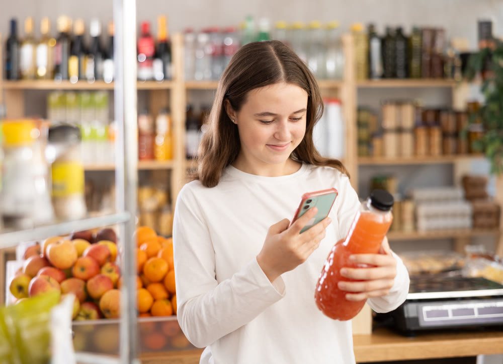 European teen girl customer at shop scans QR code on grapefruit juice bottle using phone camera