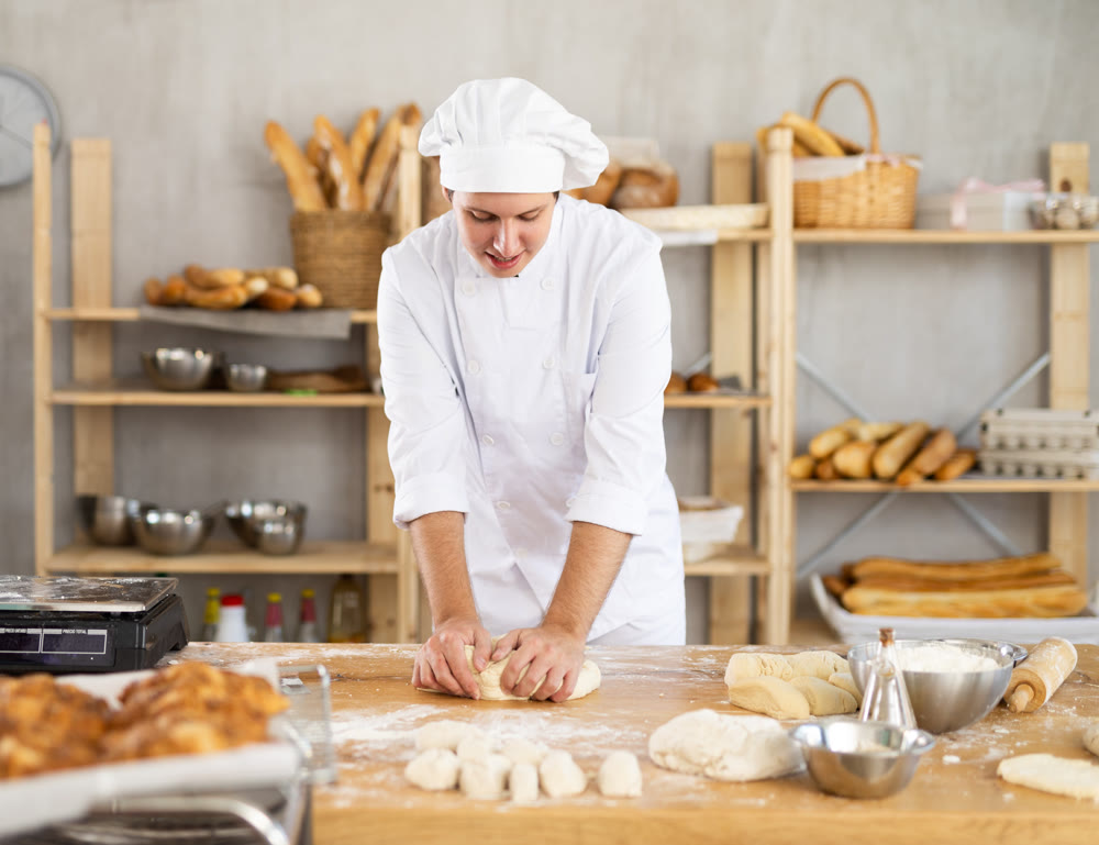 Experienced handsome young man baker in white uniform kneading dough on table in bakehouse or bakery