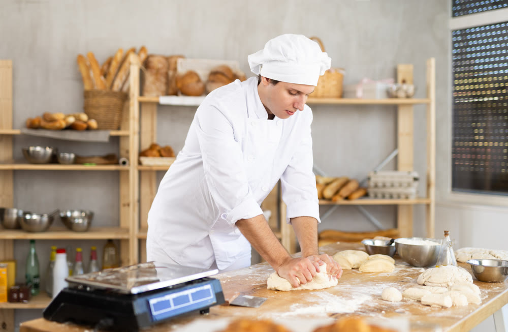 Guy baker in uniform prepares raw dough, kneads it with hands