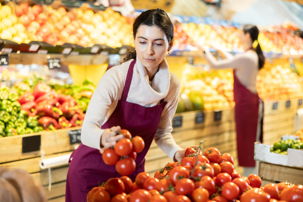 Young woman seller puts tomatoes in vegetable shop