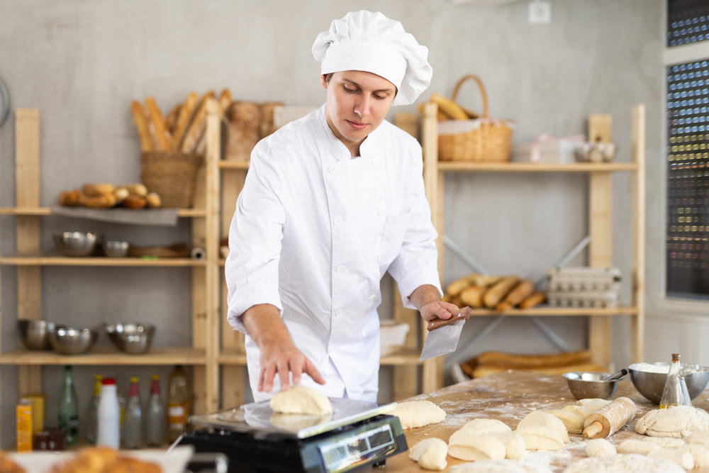 Experienced young man baker in white uniform cutting dough into pieces and weighing for baking bread on table in bakehouse or bakery