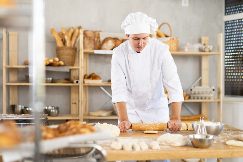 Young confidence chef guy in white uniform holding rolling pin in hands and rolling out dough in bakery kitchen