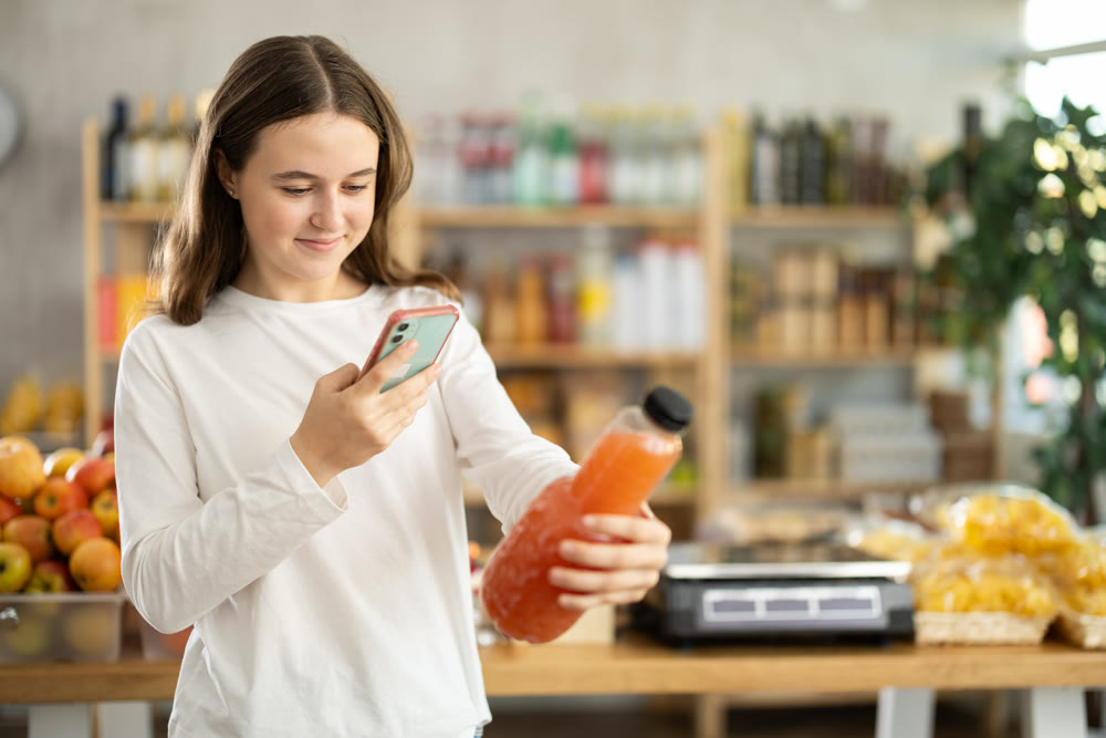 European teen girl customer at shop scans QR code on grapefruit juice bottle using phone camera