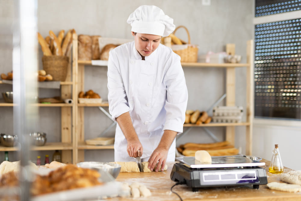 Focused male baker in white uniform working at bakery, cutting dough into pieces