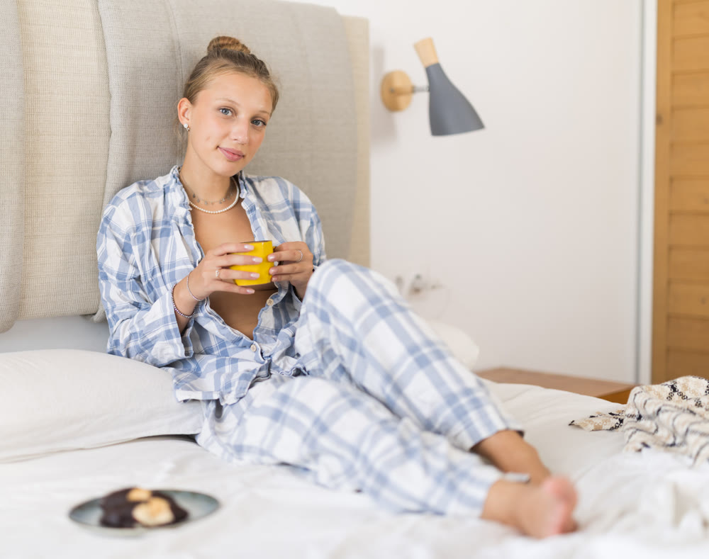 Young girl in pajamas drinking hot coffee and eating cookies while sitting on bed in bedroom - breakfast in bed