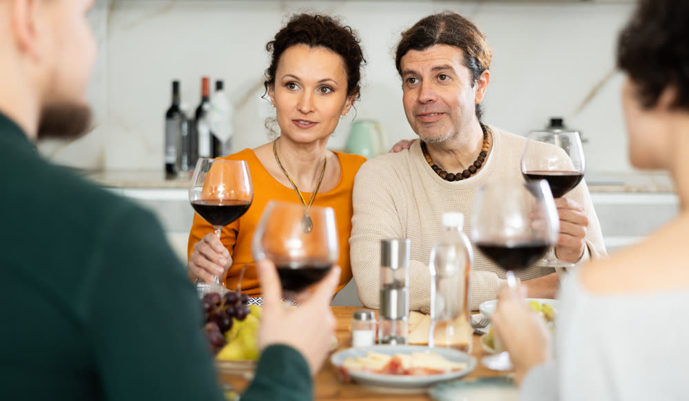 Two married couples are sitting at table, enjoying food and wine, chatting, talking, laughing.