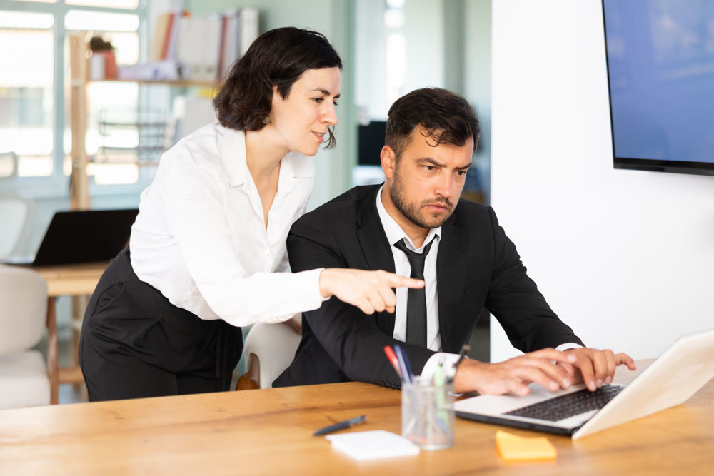 Adult woman helping adult male colleague work on laptop