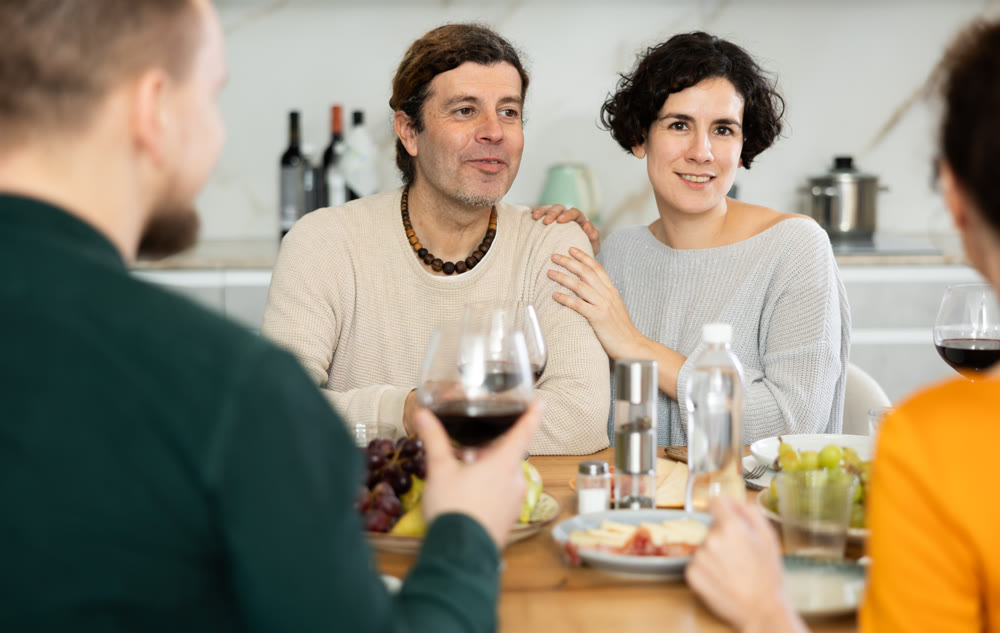 Two married couples are sitting at table, enjoying food and wine, chatting, talking, laughing.