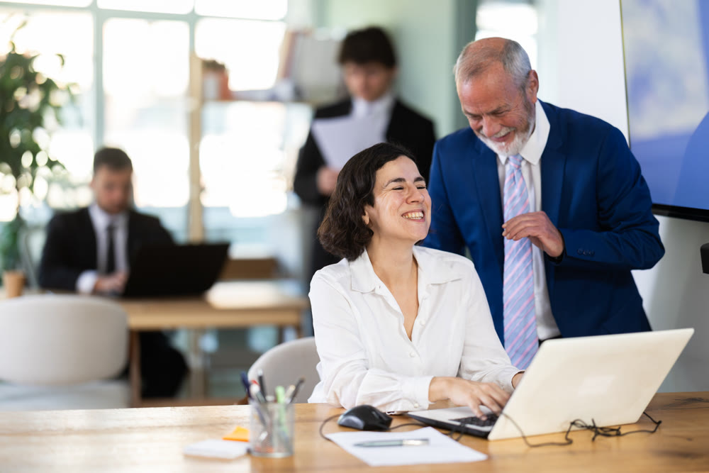 Mature male boss helping new young female business worker with laptop, discussing new project in office