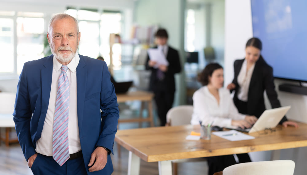 Positive mature business man in business clothes standing against background of colleague working in corporate office