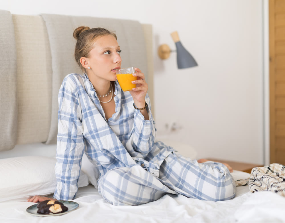 Breakfast in bed - young girl in pajamas drinking juice and eating cookies while sitting on bed in bedroom