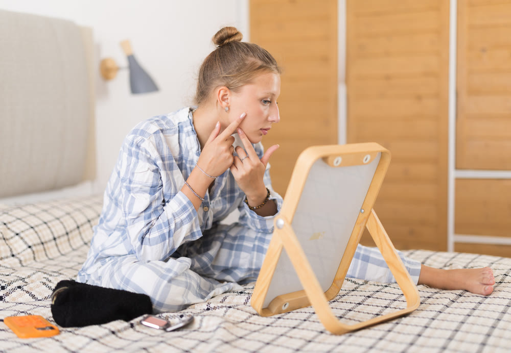 Young girl in pajamas looks in the mirror and preens herself in interior of on bed in bedroom