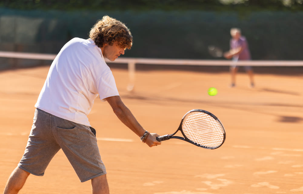 Back view of active male player hitting ball with racket on tennis court on blurred background of opponents