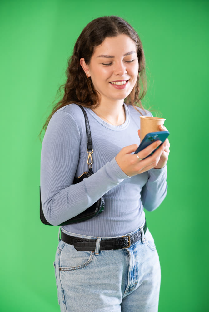 Young woman scrolling smartphone and drinking coffee in front of green screen