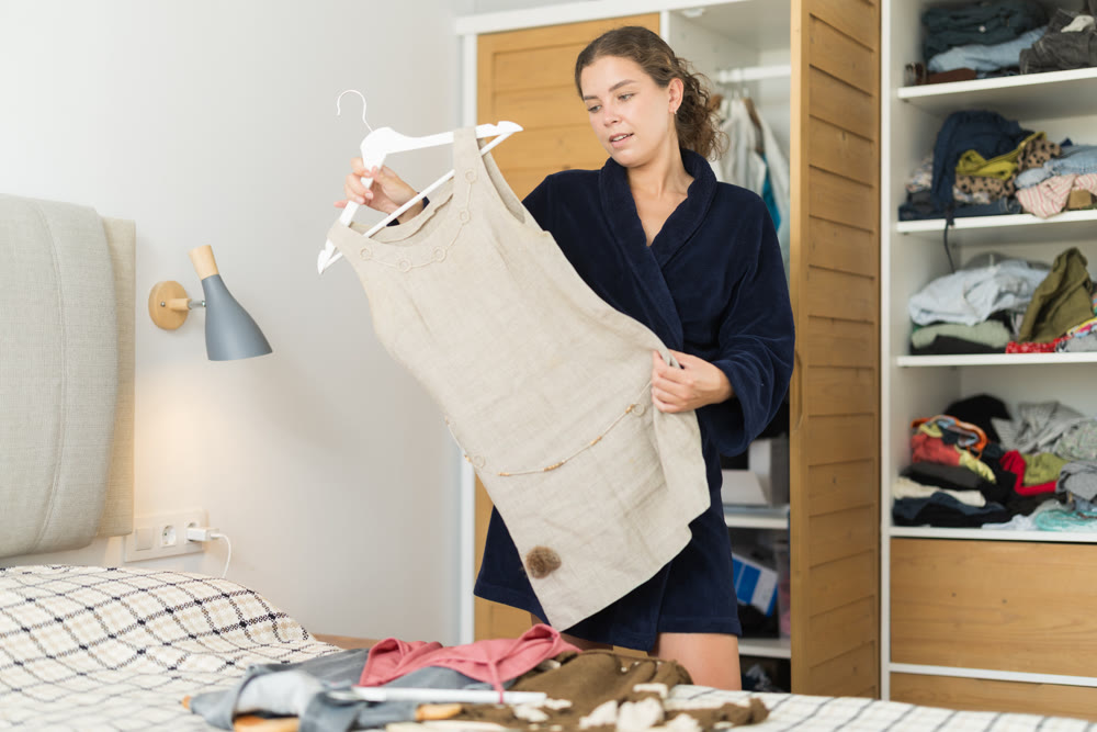 Positive young woman dressed fluffy terry-cloth bathrobe early in morning picking dress to wear for occasion in bedroom at home