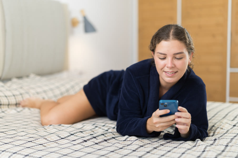 Woman in bathrobe resting in bedroom and scrolling through social media on mobile