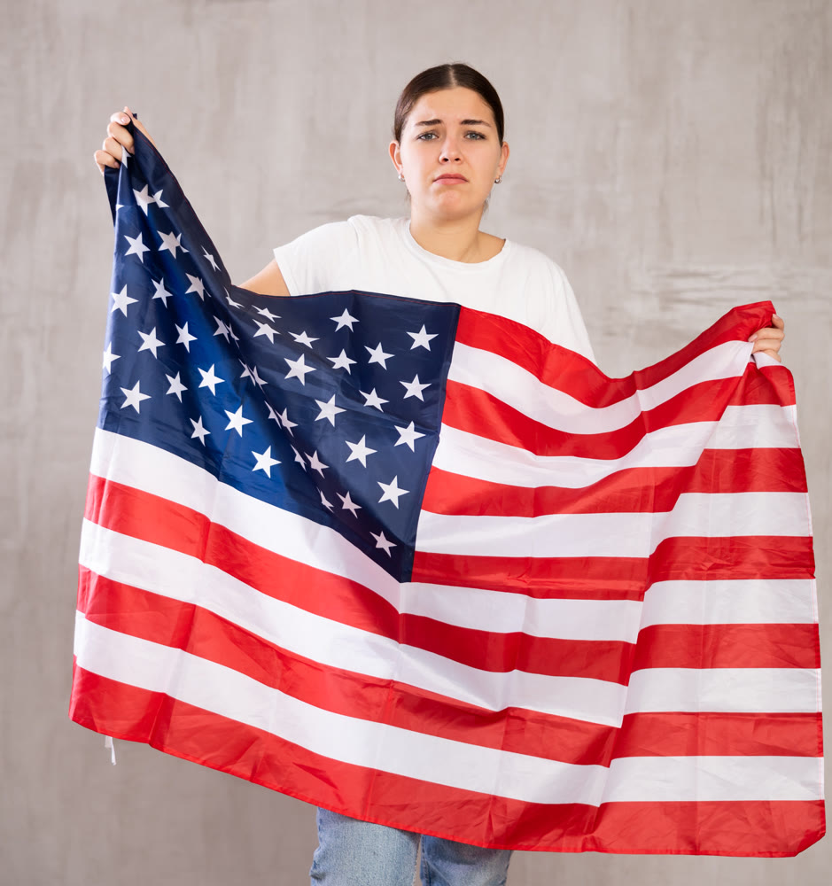Depressed young female student in casual clothes with state flag of USA against gray wall indoors