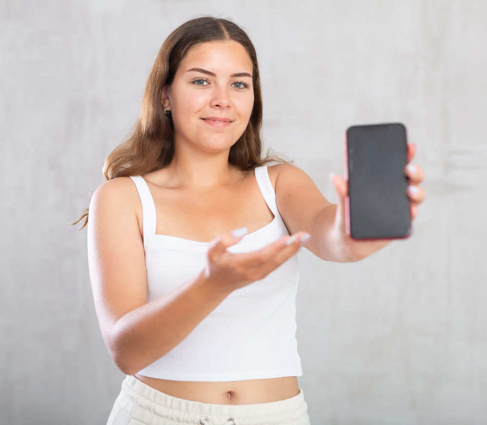 Happy young woman in casual clothes showing screen of her smartphone